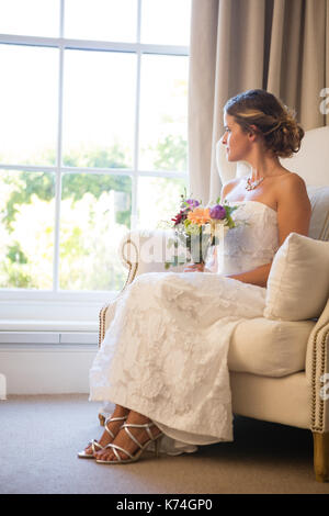 Belle bride holding bouquet à la fenêtre à travers tout en restant assis sur un fauteuil à la maison Banque D'Images