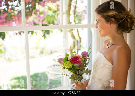 Vue latérale du smiling beautiful bride holding bouquet tout en regardant par la fenêtre à la maison Banque D'Images