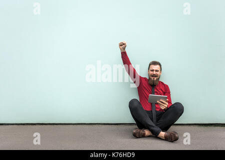 Travaillant en plein air, se trouve sur le terrain. jeune homme barbu adultes travaillent en comprimé, lève la main et crier avec sourire. Piscine tourné sur mur gris. Banque D'Images