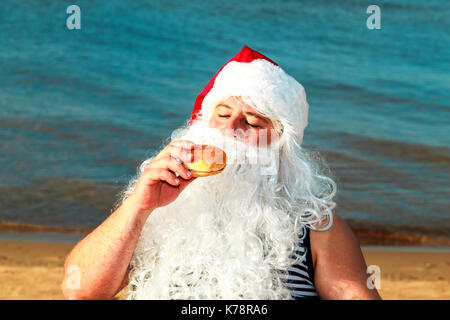 Père Noël sur la plage de manger un hamburger. Le concept de l'alimentation malsaine. Banque D'Images