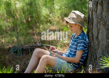 Boy leaning against tree trunk, using digital tablet Banque D'Images