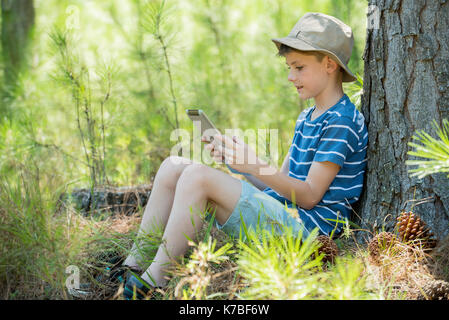 Boy leaning against tree trunk, using digital tablet Banque D'Images