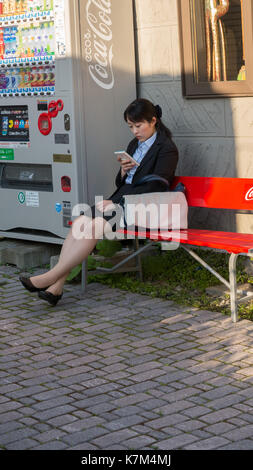 Jeune femme japonaise en faisant une pause assis sur la banquette rouge en costume noir et chemise bleue à la téléphonie cellulaire et la luminosité de l'après-midi chaud. Banque D'Images