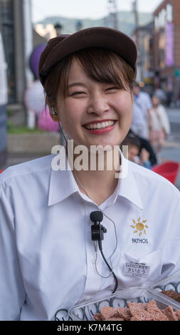 Young smiling woman touristes japonais des échantillons de pathos cookies en face du magasin. light blue shirt, badge, le pathos des lèvres rouges et des dents blanches logo Banque D'Images