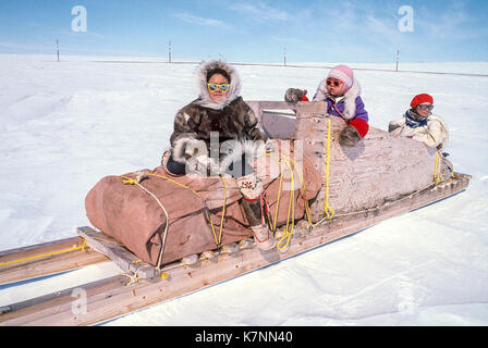 Les enfants inuits, les filles âgées de 11 et 4, vêtu de vêtements traditionnels en peau de caribou, avec femme ancien, assis sur un traîneau de fret traditionnel des Inuits, également appelé un traîneau. Banque D'Images