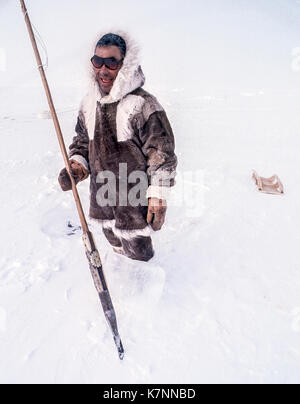 Aîné inuit homme, milieu des années 60, vêtu de vêtements traditionnels en peau de caribou, pose avec l'outil qu'il utilise pour couper des blocs de glace pour construire des igloos. Banque D'Images