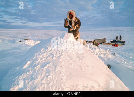 Aîné inuit homme, milieu des années 60, vêtu de vêtements traditionnels en peau de caribou, travaille sur un igloo traditionnel avec un couteau utilisé pour la construction d'igloos. igloos réelles ne sont pas en reste avec des blocs montrant. quand les blocs sont en place, l'igloo est recouvert de neige pour plus d'isolement. un traîneau traditionnel (traîneau) peut être vu en arrière-plan. Banque D'Images