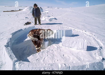 Aîné inuit homme vêtu de vêtements traditionnels en peau de caribou, construit un igloo en sculptant des blocs de neige et de les placer avec soin. c'est un abri traditionnel toujours utilisé par certains Inuits aujourd'hui. un autre aîné inuit homme, vêtu de vêtements de l'Arctique, les montres modernes. Banque D'Images
