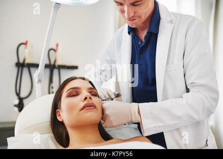 Médecin homme d'effectuer des injections de botox à la joue d'une jeune femme allongée sur une table dans une clinique de beauté Banque D'Images