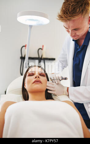 Jeune homme médecin d'effectuer des injections de botox à la joue d'une femme allongée sur une table dans une clinique de beauté Banque D'Images