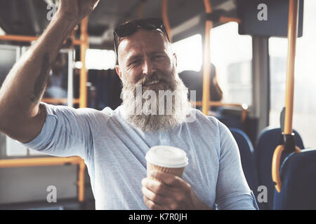 Smiling mature man avec une longue barbe et de boire une tasse de café à emporter en étant debout sur un bus pendant son trajet du matin Banque D'Images
