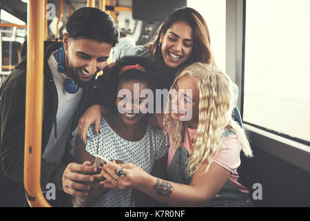 Groupe diversifié de smiling friends sitting sur un bus de médias sociaux ensemble sur un smartphone Banque D'Images
