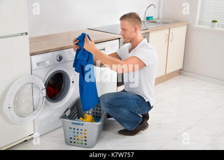 Jeune homme à la recherche de t-shirt lors de l'utilisation de lave-linge appareil dans la cuisine Banque D'Images