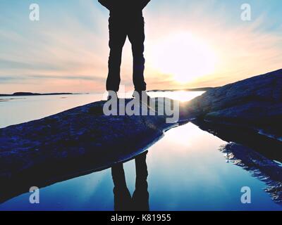 Grand randonneur dans dark sportswear avec sac à dos sportif se dresse sur falaise au-dessus de la mer. L'homme profitez incroyable coucher du soleil à l'horizon avec orange ciel rose avec des nuages. ma Banque D'Images