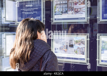 Une jeune femme à la recherche de maisons inabordables à vendre et à louer dans une fenêtre d'agents immobiliers à Londres, Royaume-Uni. Banque D'Images