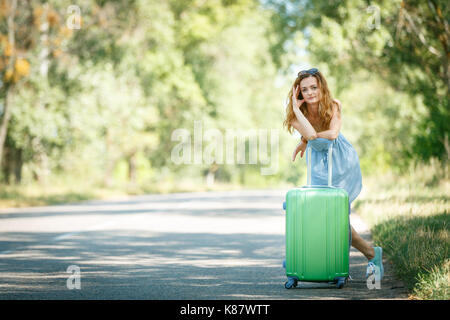 Fatigué de l'auto-stop fille dans une robe d'été s'appuyant sur un cas. Road adventure concept Banque D'Images