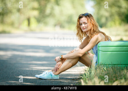 Fatigué de l'auto-stop fille dans une robe d'été assis sur une route avec un boîtier en plastique vert. Road adventure concept Banque D'Images