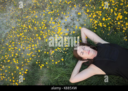 Une femme dans un débardeur noir et un jean couché dans l'herbe avec ses mains derrière sa tête. Banque D'Images