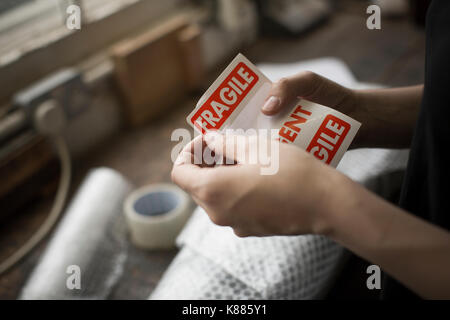 Mains tenant un fragile rouge autocollant à coller sur un paquet de papier brun sur un établi. Banque D'Images