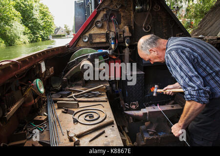 Forgeron travaillant dans un petit espace sur son grand classique, une barge sur la rivière, penché au-dessus de l'enclume et façonner le métal chaud. Banque D'Images