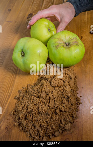 A woman's hand holding l'une des trois matières, Bramley, aux côtés de pommes à cuire le sucre brun doux sur un plan de travail de cuisine pin, prêt pour la cuisson. Angleterre, Royaume-Uni. Banque D'Images