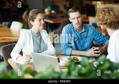 Les jeunes gestionnaires talentueux brainstorming sur projet prometteur et prendre les notes pendant que se sont réunis au restaurant moderne, arrière-plan flou Banque D'Images