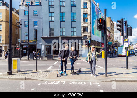 Couple sur les scooters de poussée attendant de traverser la route à Shoreditch, Londres, Royaume-Uni Banque D'Images