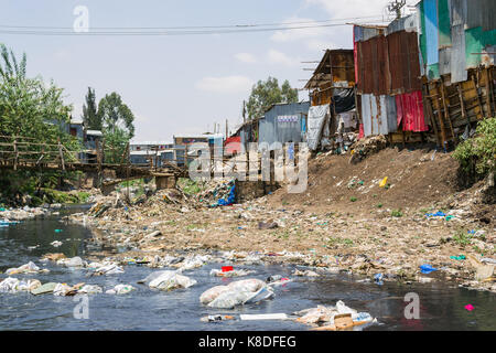 La ligne des baraques taudis rivière Ngong qui est polluée par des déchets, les déchets plastiques et les déchets, un pont en bois de fortune le traverse, Nairobi, Kenya Banque D'Images