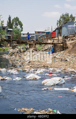 La ligne des baraques taudis rivière Ngong qui est polluée par des déchets, les déchets plastiques et les déchets, un pont en bois de fortune le traverse, Nairobi, Kenya Banque D'Images