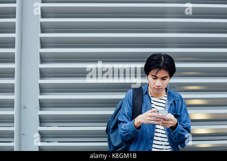 Jeune homme se tenant debout à l'extérieur, à l'aide de smartphone, wearing earphones Banque D'Images