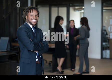 Portrait of businessman, bras croisés, sourire, businesswomen talking in background Banque D'Images