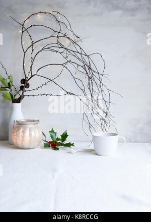 Un arrangement de Noël simple avec des branches, des brindilles, des lumières de fées, des cônes de pin et des houx, une bougie allumée, et un teacup. Style photo, lumière naturelle Banque D'Images