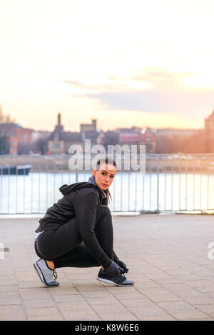 Une belle jeune femme attachant sa chaussure dans le coucher du soleil au bord de la rivière tandis que crouching Banque D'Images