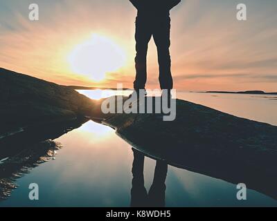 Randonneur dans l'homme les jambes hautes bottes en miroir dans l'eau de mer chauffée, avec coucher de soleil.'figure debout sur piton rocheux. rayons de soleil faire reflet dans l'eau salée Banque D'Images