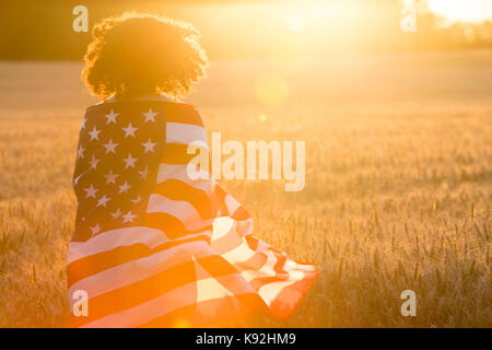 Mixed Race african american girl adolescent femme jeune femme dans un champ de blé ou d'orge enveloppés dans usa stars and stripes flag dans golden sunse Banque D'Images