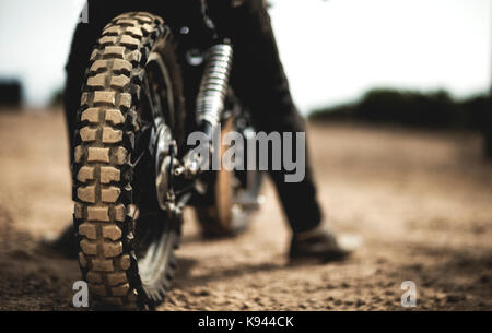 Vue arrière de l'homme assis sur le cafe racer moto sur un chemin de terre poussiéreux, Close up de pneu. Banque D'Images