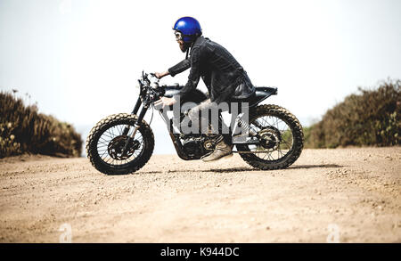 Vue de côté d'un homme portant un casque bleu face ouverte et lunettes équitation cafe racer moto sur une route de terre poussiéreuse. Banque D'Images