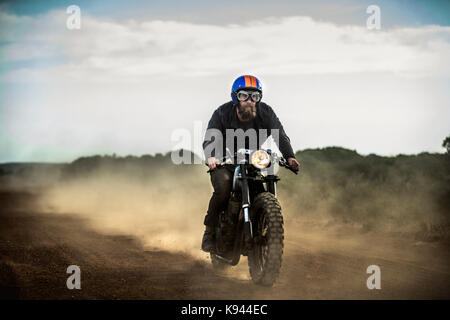 Face ouverte homme portant un casque et lunettes équitation cafe racer moto sur une route de terre poussiéreuse. Banque D'Images