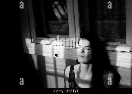 Jeune femme se dorer sous les rayons du soleil dans l'ombre de la fenêtre des trames sur le porche. la photographie noir et blanc. Banque D'Images