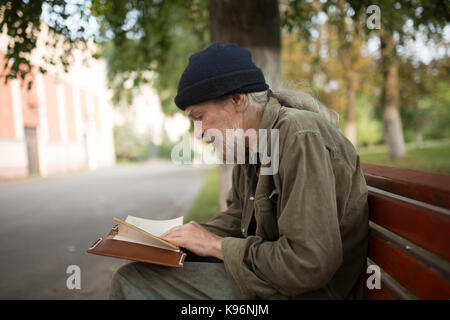 Vue latérale du vieux sans-abri avec de longs cheveux gris en lisant un livre. Banque D'Images