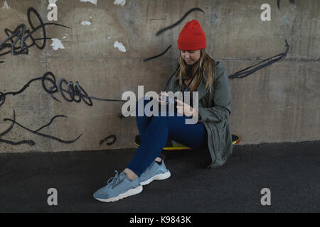 Young woman using mobile phone while sitting on skateboard contre mur Banque D'Images