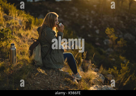 Female hiker ayant un verre tout en étant assis sur la montagne au coucher du soleil Banque D'Images