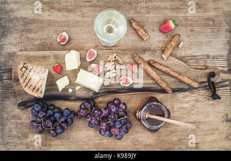Verre de vin blanc, plateau de fromages, raisins, figues, fraises, miel et gressins sur fond de bois rustique Banque D'Images