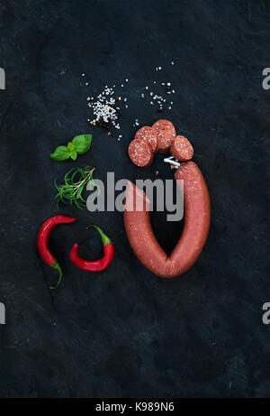 Salami fumé saucisse avec le romarin, le piment, le basilic et le sel noir sur fond noir en ardoise, vue du dessus. Banque D'Images