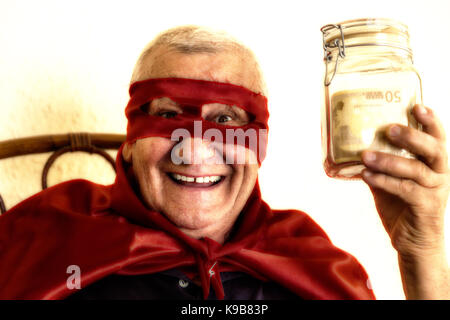 Vieil homme habillé en super héros, en souriant, assis sur chaise en osier et tenant un pot plein de billets sur fond de mur jaune Banque D'Images