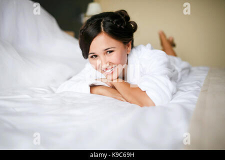 Mariée joyeuse le matin sur le lit en peignoir smiling joyeuse, positive, avec un prêt la coiffure et maquillage, une robe de mariage dans l'arrière-plan. En prévision du mariage. Banque D'Images