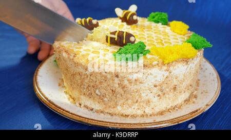 Woman's hands couper le gâteau à la crème de miel sur fond bleu. Gâteau au miel. gâteau avec les abeilles. coupe le cake Banque D'Images