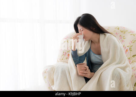 Jolie femme maladie attraper froid à l'aide de nez de soufflage de tissus et de boire de l'eau chaude chambre assis sur fauteuil relaxant. Banque D'Images