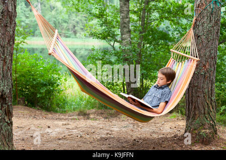 Garçon couché dans un hamac en lisant un livre à l'extérieur Banque D'Images