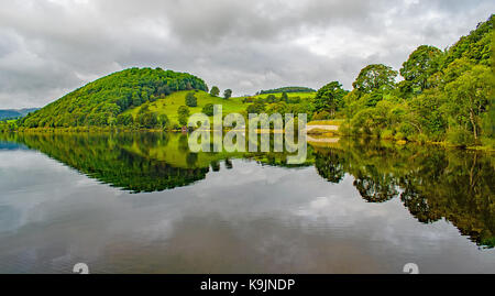 Lake Ullswater's green Rolling hills reflète la symétrie est contre un ciel maussade. Banque D'Images